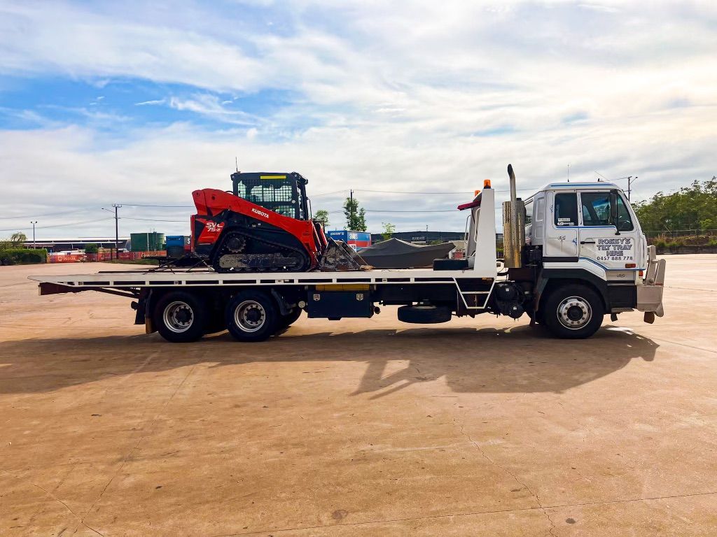 A Flatbed Truck With a Tractor on the Back is Parked in a Dirt Lot — Fewmore Contracting in Palmerston, NT