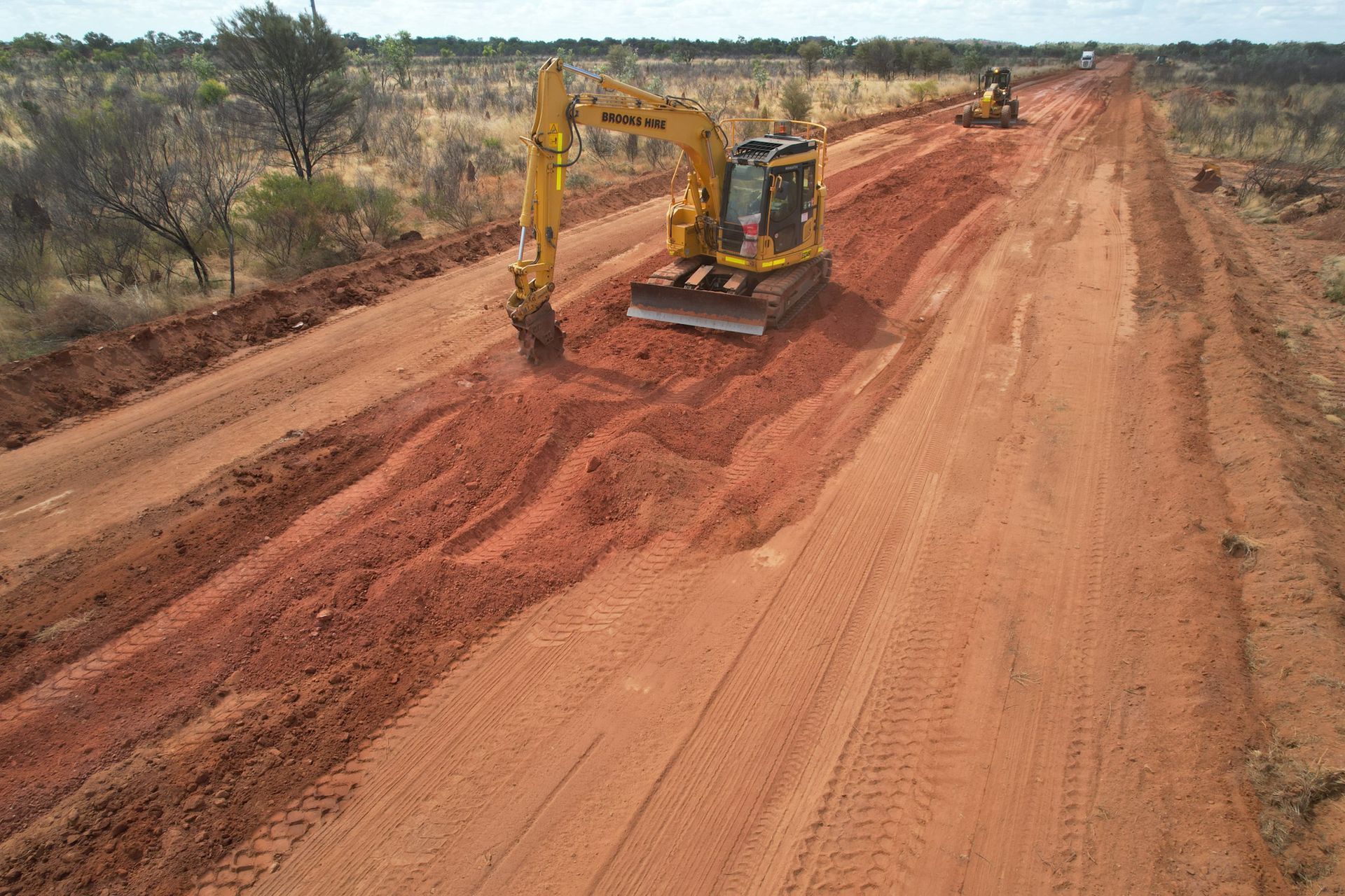 A Yellow Excavator is Driving Down a Dirt Road — Fewmore Contracting in Katherine, NT