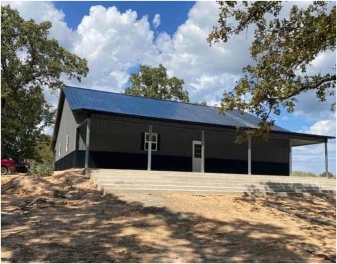 A grey barn-style building with a blue metal roof and a large concrete patio, surrounded by trees under a cloudy sky.