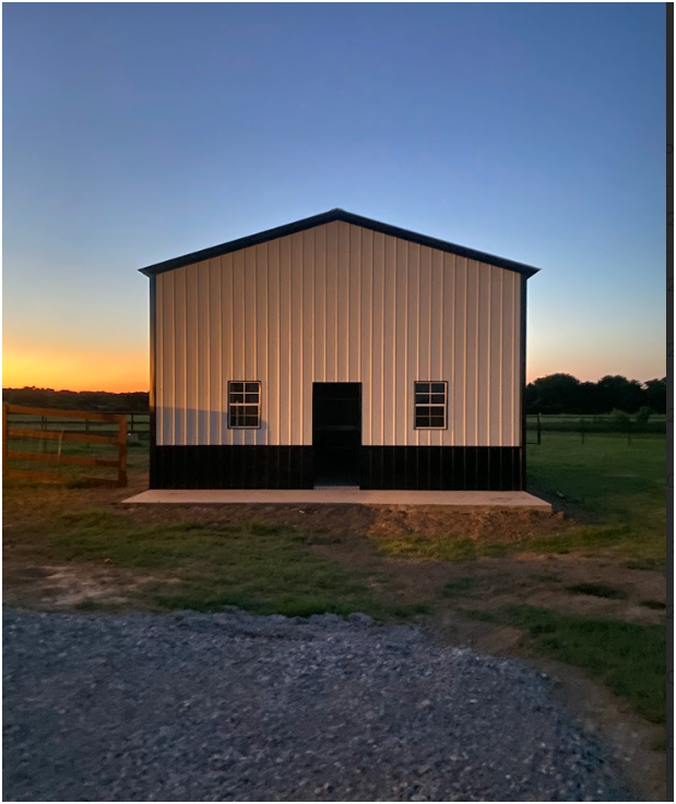A tan metal building with black trim, a center door, and two windows, set in a field during sunset.