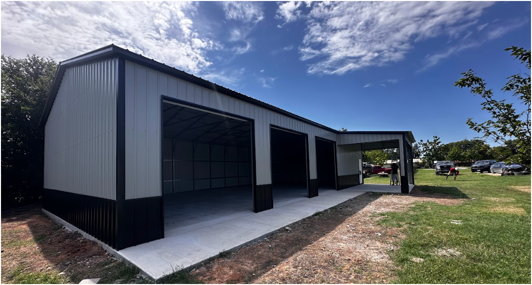 A gray metal garage with a black trim and base, three open bays, and a side carport on a grassy lot under a blue sky.