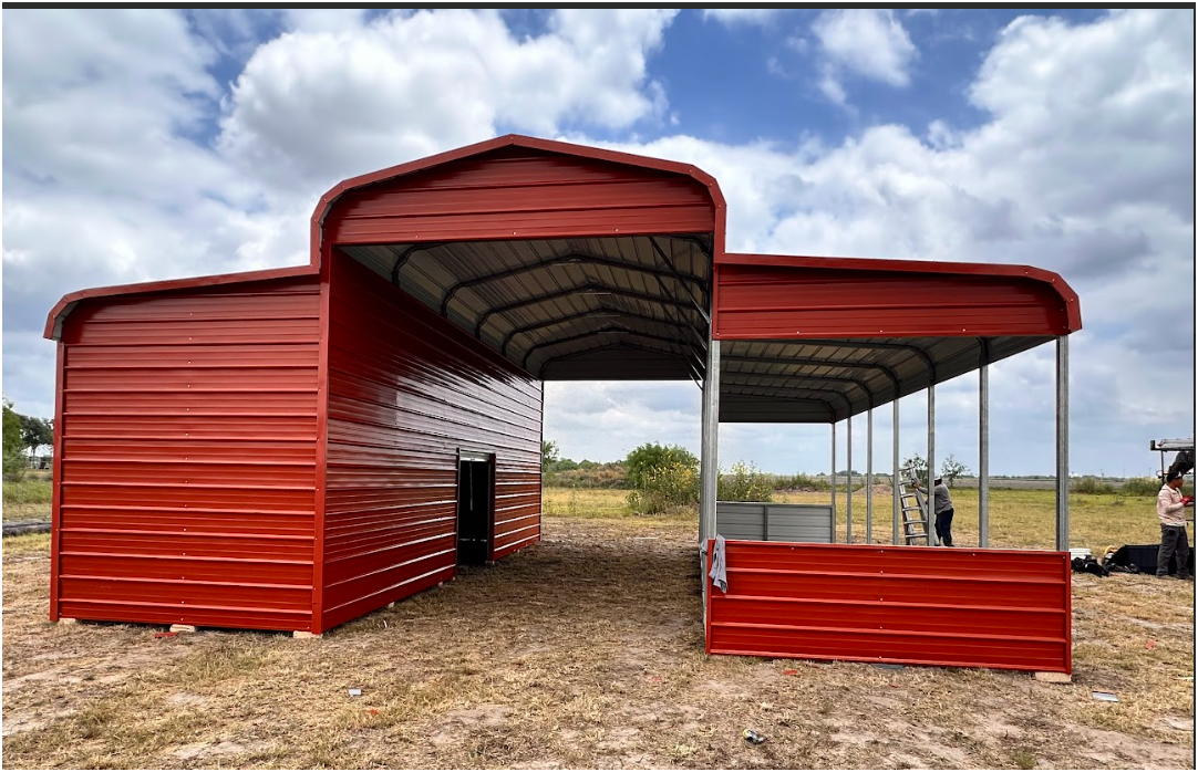 A red metal barn structure with a center raised roof section and an open-sided lean-to, standing in a grassy field.