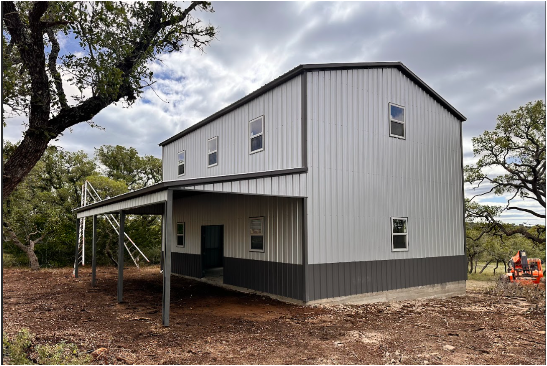 A two-story metal barn-style house with gray and white siding, featuring a covered porch, set in a wooded landscape.