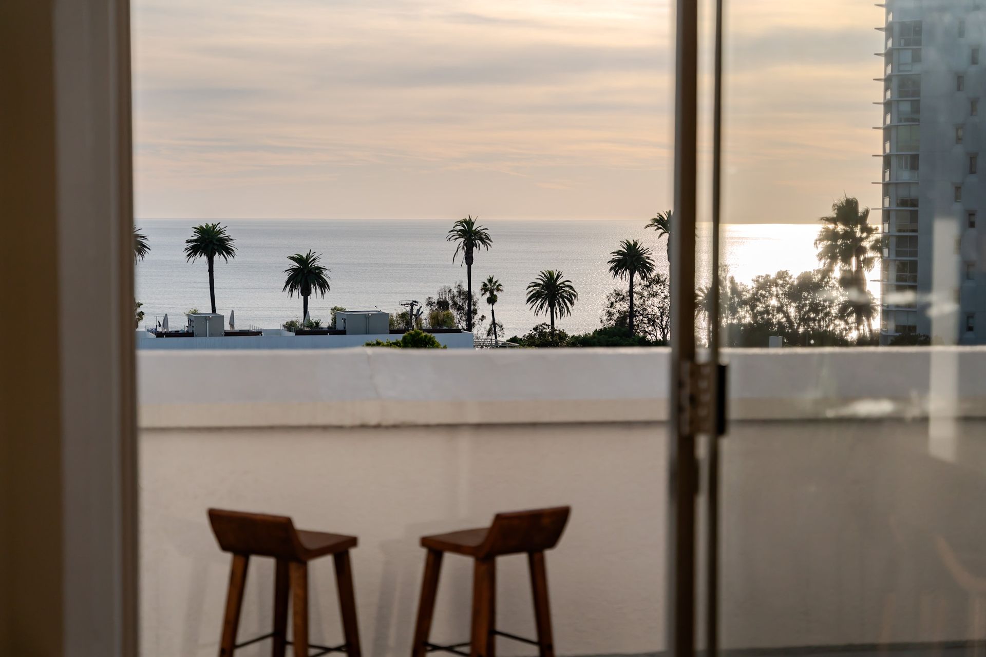 View from balcony with two wooden stools overlooking ocean, palm trees, and building.