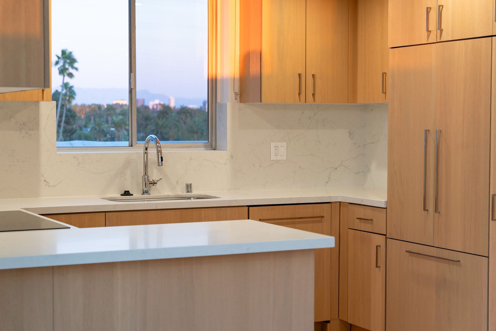 Modern kitchen with light wood cabinets, white countertops, and a window with an outside view.