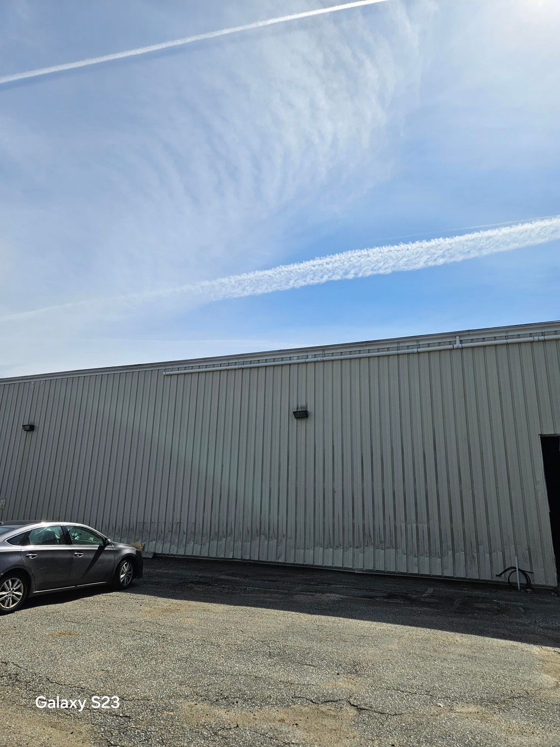 Gray building with a wavy side, car parked on gravel, blue sky with contrails.