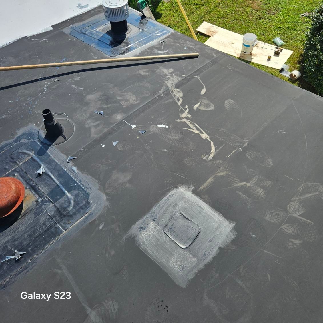 Black rooftop with vents, flashing, and patch repairs near a grassy yard.