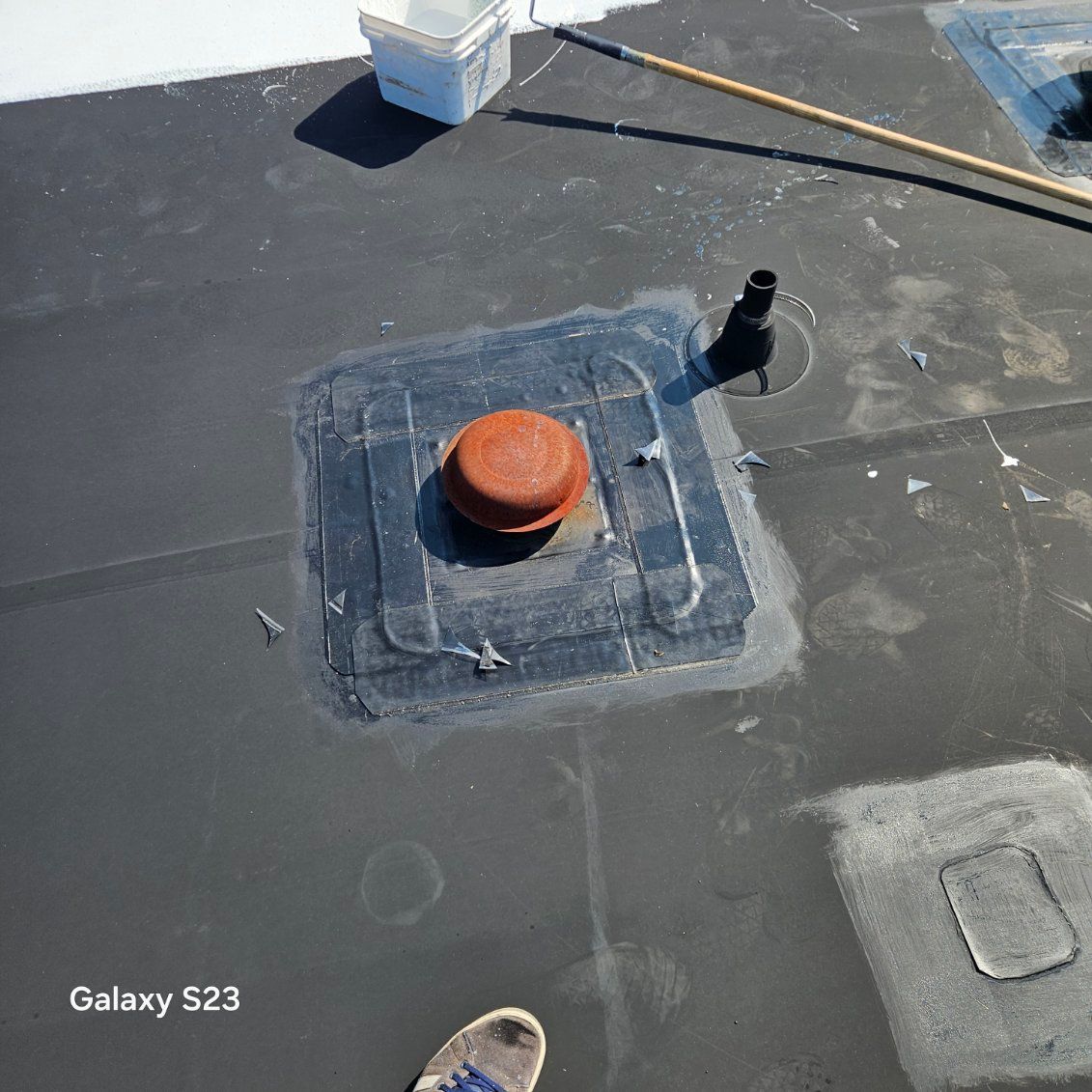 Rooftop view of a black membrane roof with vent pipes, a red cap, and a small white roof unit.