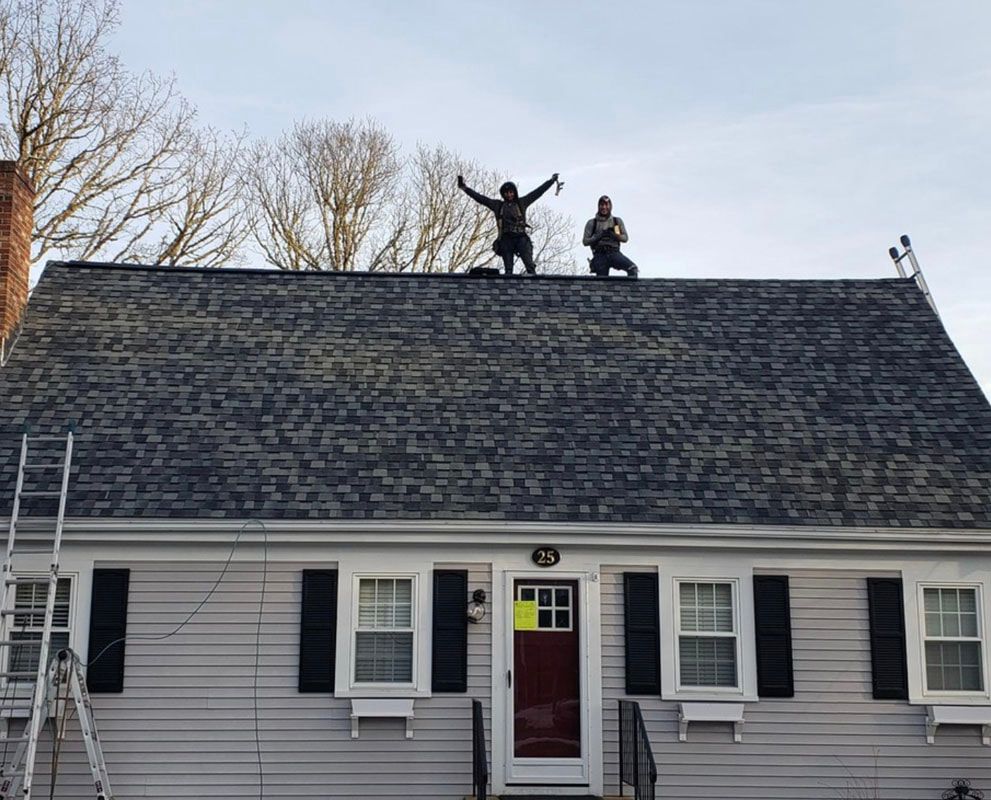 Two roofers on a gray shingled roof; one with arms raised.  A gray house with black shutters.