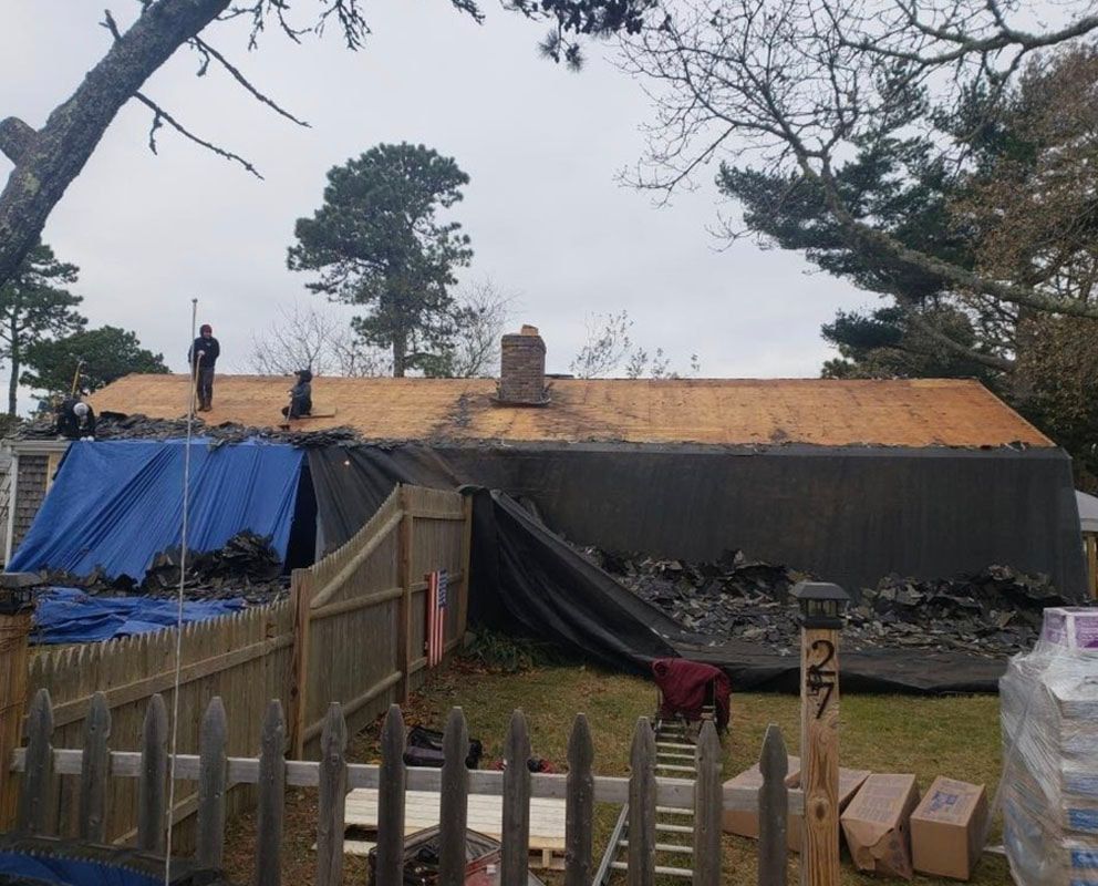 Roofers working on a house, covered with tarps. Blue tarp in front of the roof and a brown wooden fence.