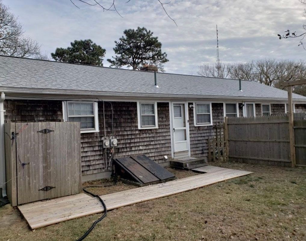 A weathered, one-story cedar shingle house with a wooden shed, platform, and fence in the backyard.