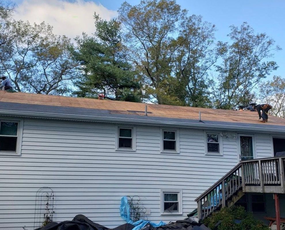 Workers on a rooftop, installing shingles. White siding, trees in background, and a wooden deck.