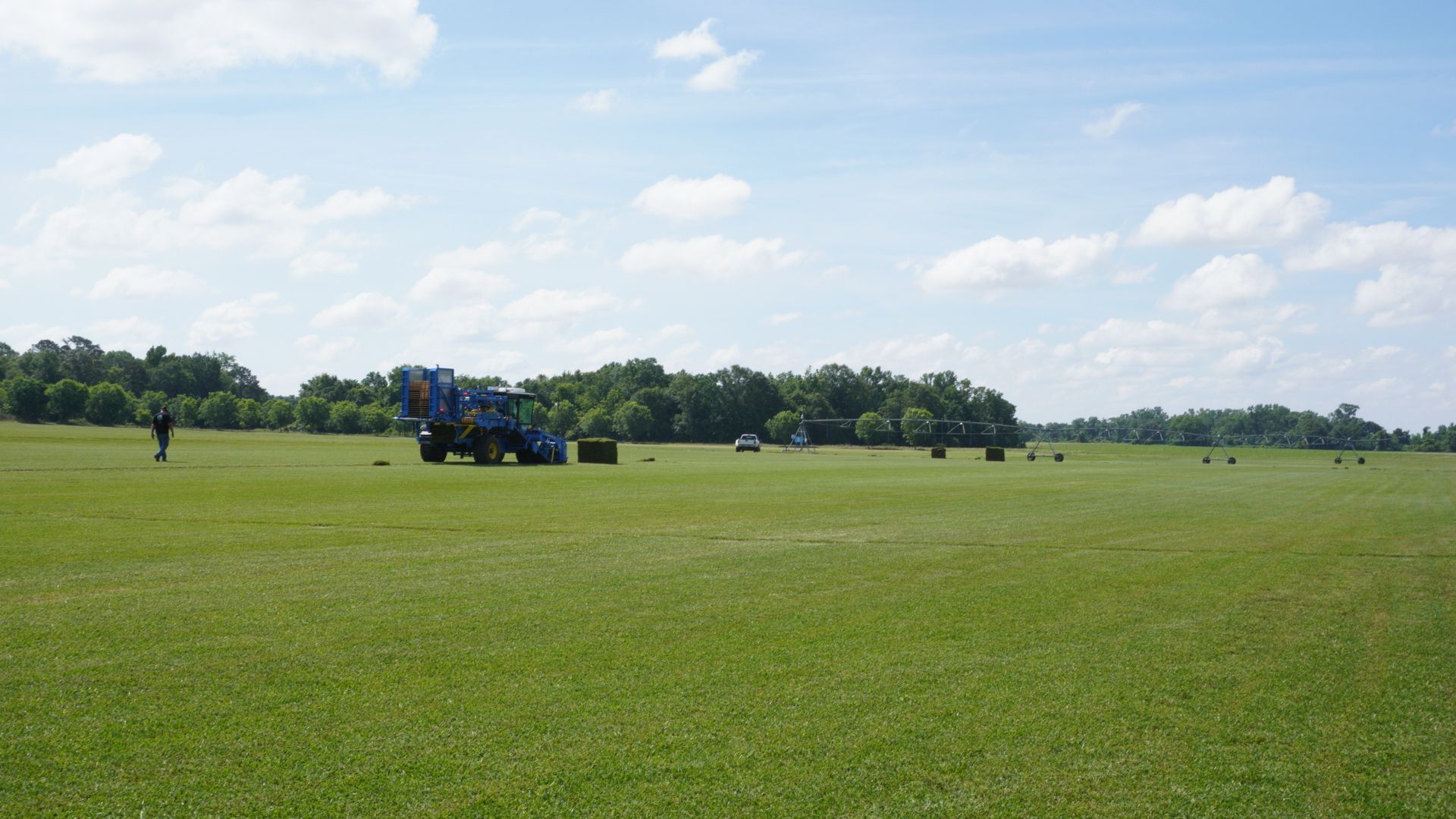 Grassy field with a blue tractor in the distance under a bright, partly cloudy sky