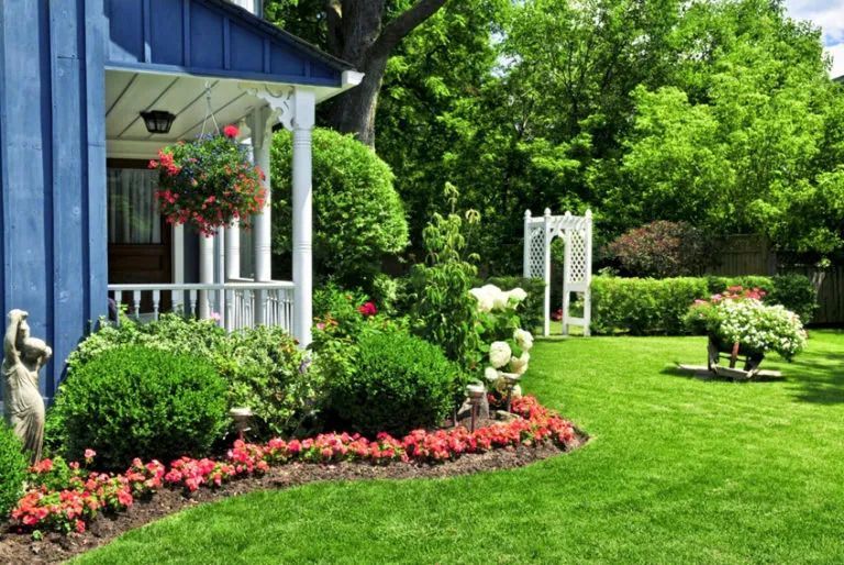 Blue house with porch and lush garden, pink flowers, white arbor, and manicured green lawn