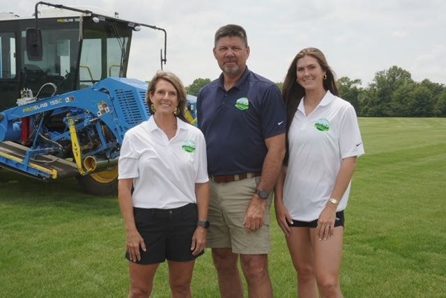 Three people posing on a grassy field beside a blue tractor and sprayer equipment
