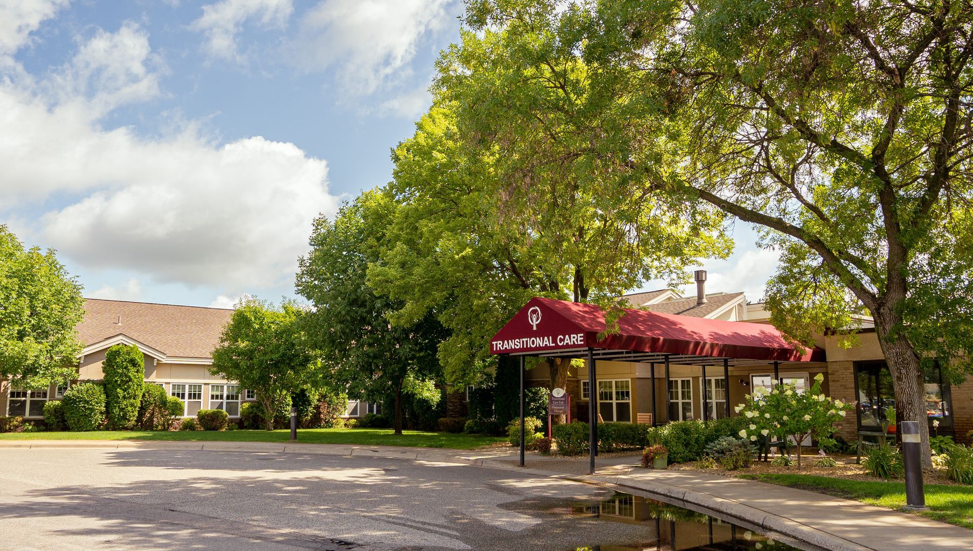A low-rise building with a red fabric awning over the entrance, surrounded by green trees under a partly cloudy sky.