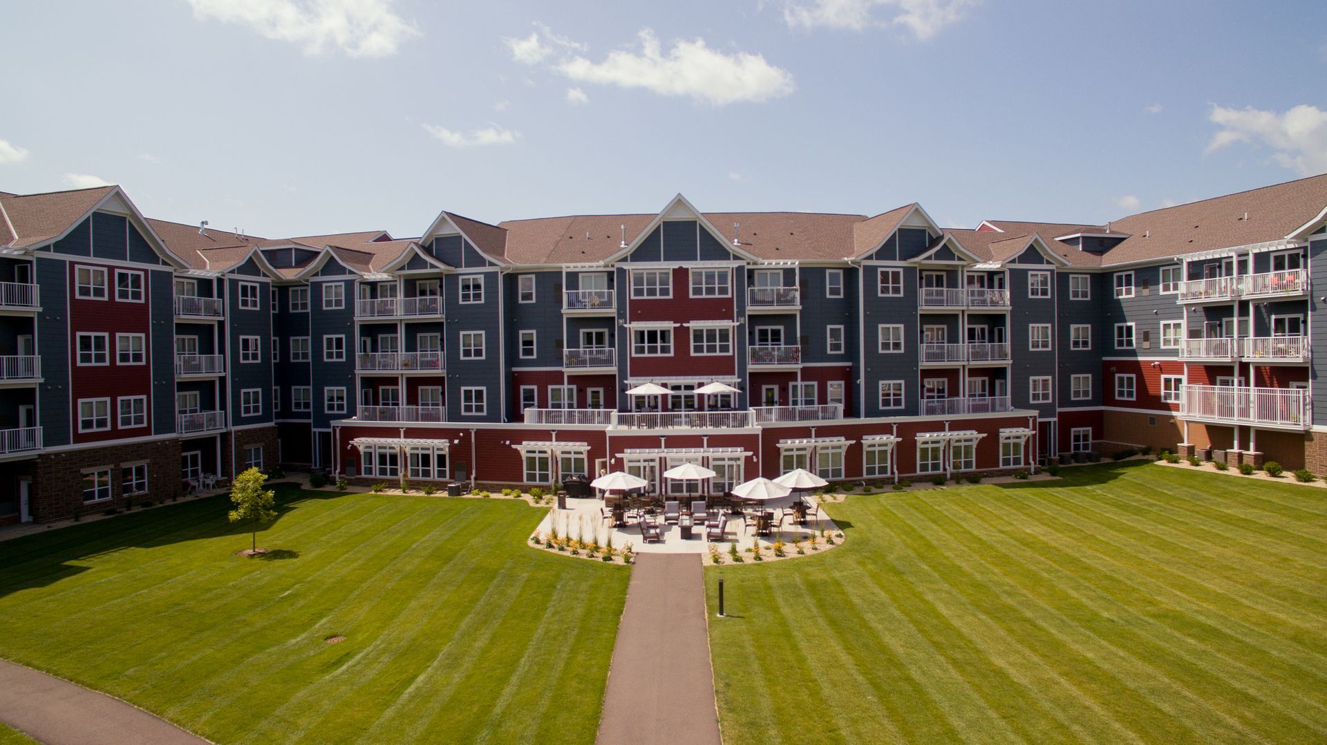 A multi-story apartment building with red and blue siding, overlooking a large green lawn with a central patio area.
