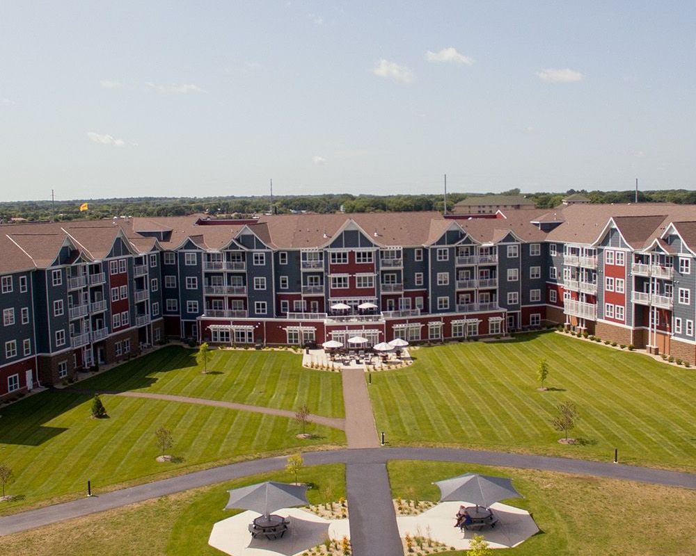An aerial view of a multi-story apartment complex with dark blue and red siding, surrounded by a large, mowed grass lawn.