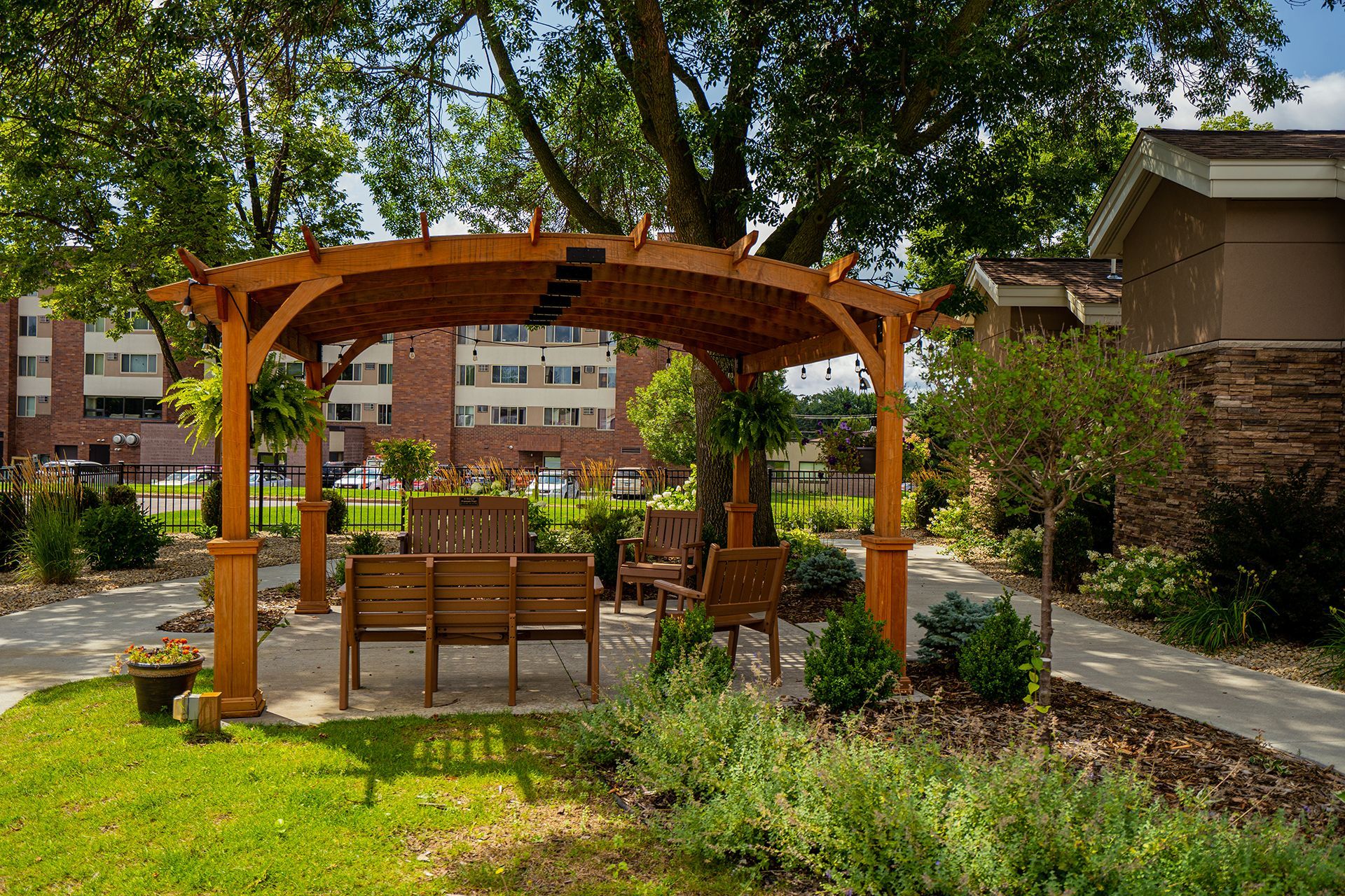 A wooden pergola shades a small seating area with benches in a sunny, landscaped garden by apartment buildings.