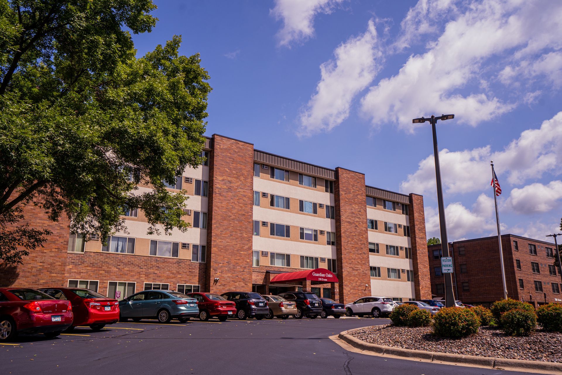 A multi-story brick hotel with a red awning entrance, surrounded by a parking lot, trees, and a flag under a blue sky.