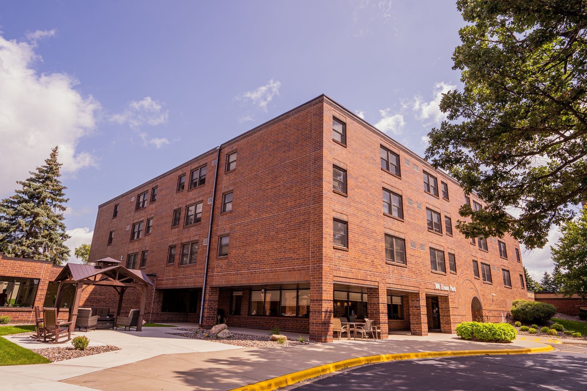A multi-story brick residential building with a patio, wooden gazebo, and surrounding trees under a bright blue sky.