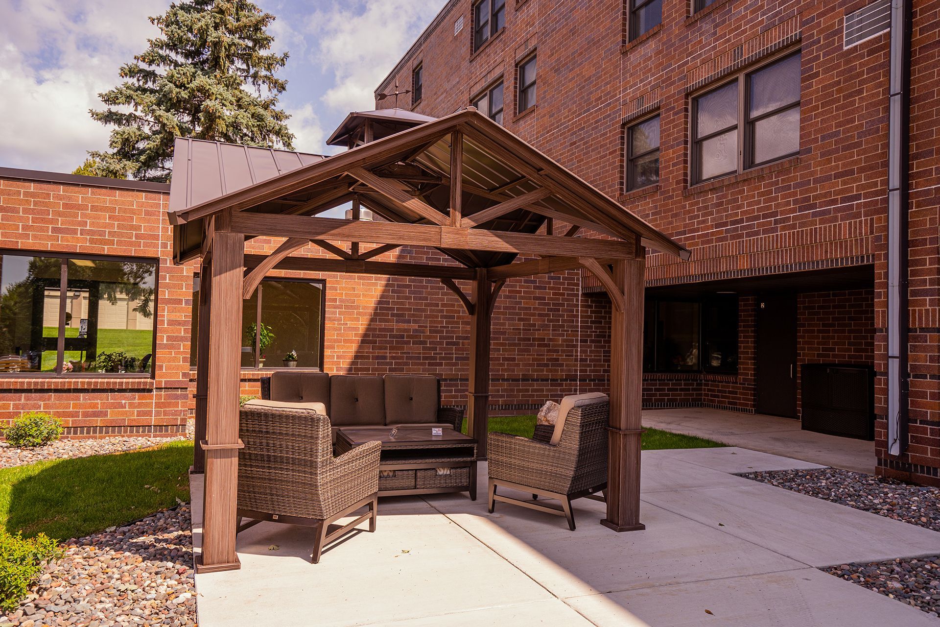 Wicker chairs and a sofa sit under a wooden gazebo on a concrete patio next to a red brick building.