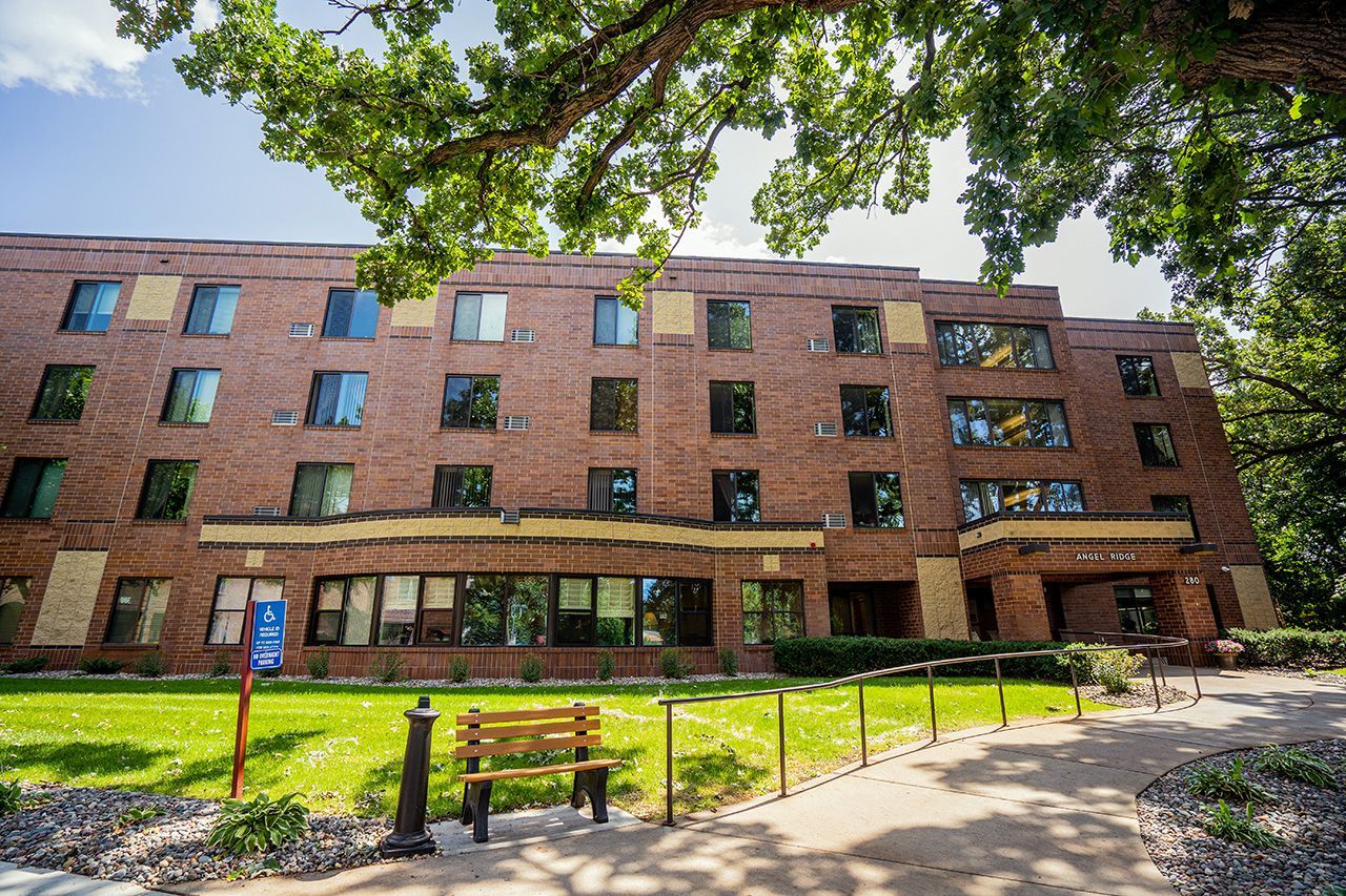 A four-story brick campus building with large windows, a front lawn, and a wooden bench under leafy green trees.