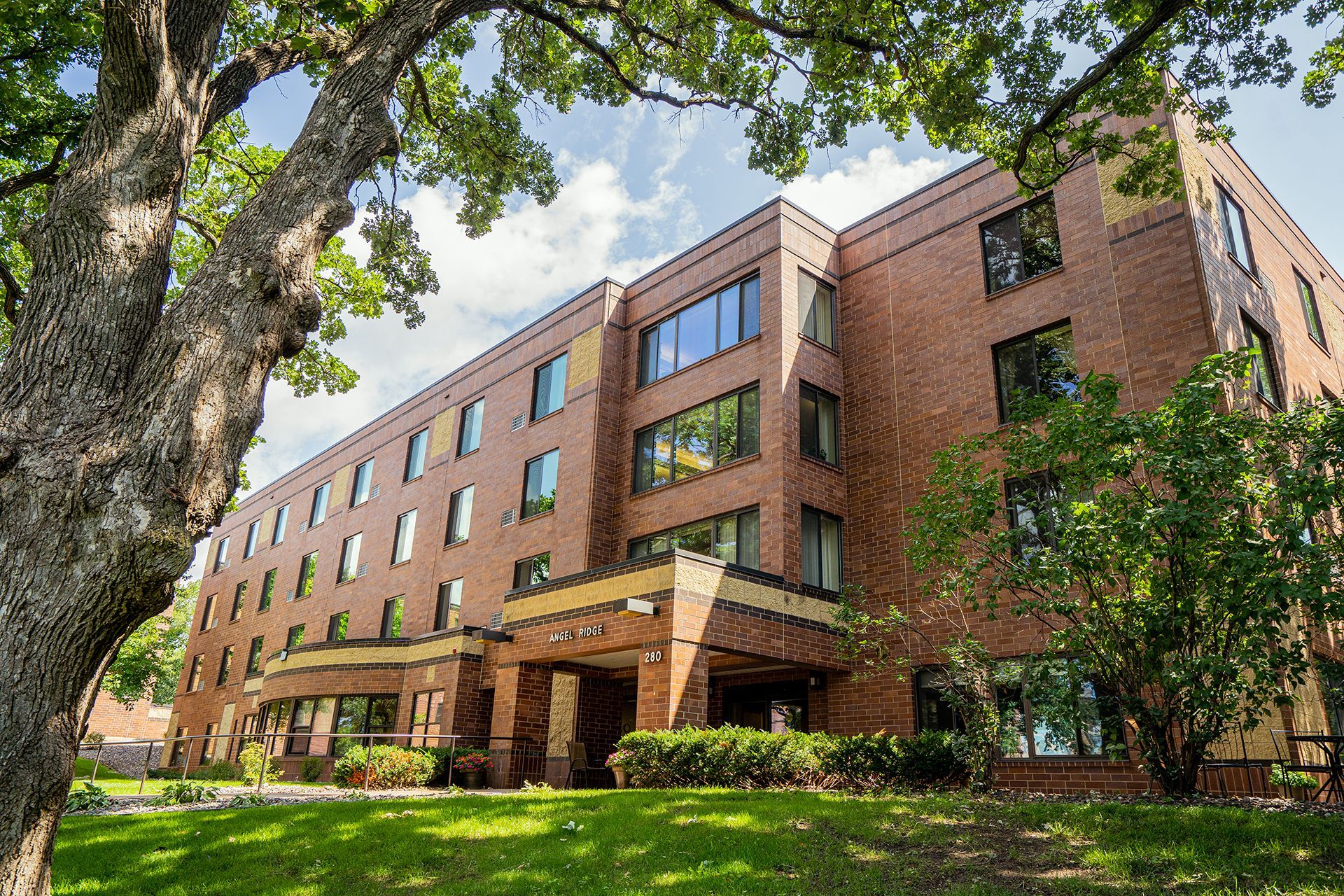 A multistory brick building with rows of windows and a ground-floor entrance, surrounded by trees and a green lawn.