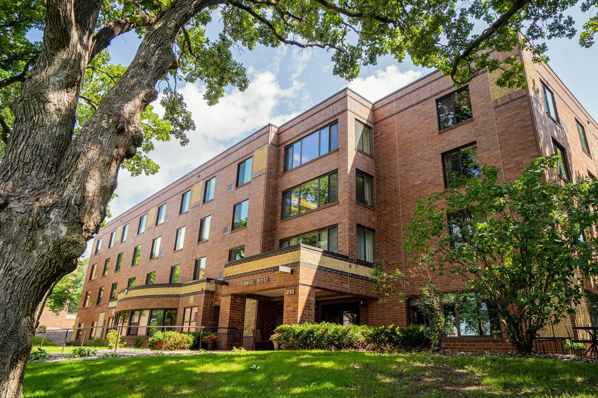 A low-angle view of a multi-story red brick building with large windows, surrounded by green trees and a grassy lawn.