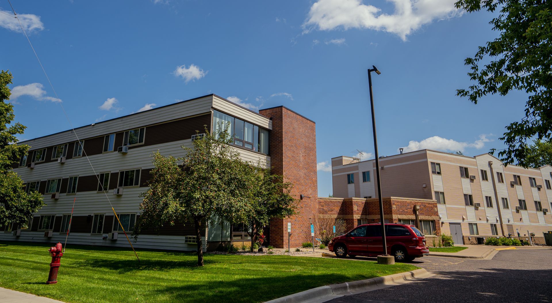 A multi-story brick and white apartment building sits on a grassy lawn under a blue sky, with a red minivan parked nearby.