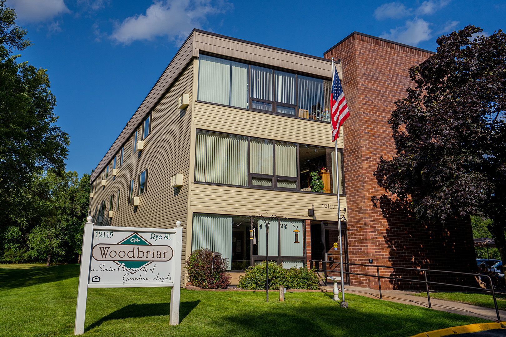 Three-story apartment building with beige siding and a brick facade, featuring a sign in front and an American flag.