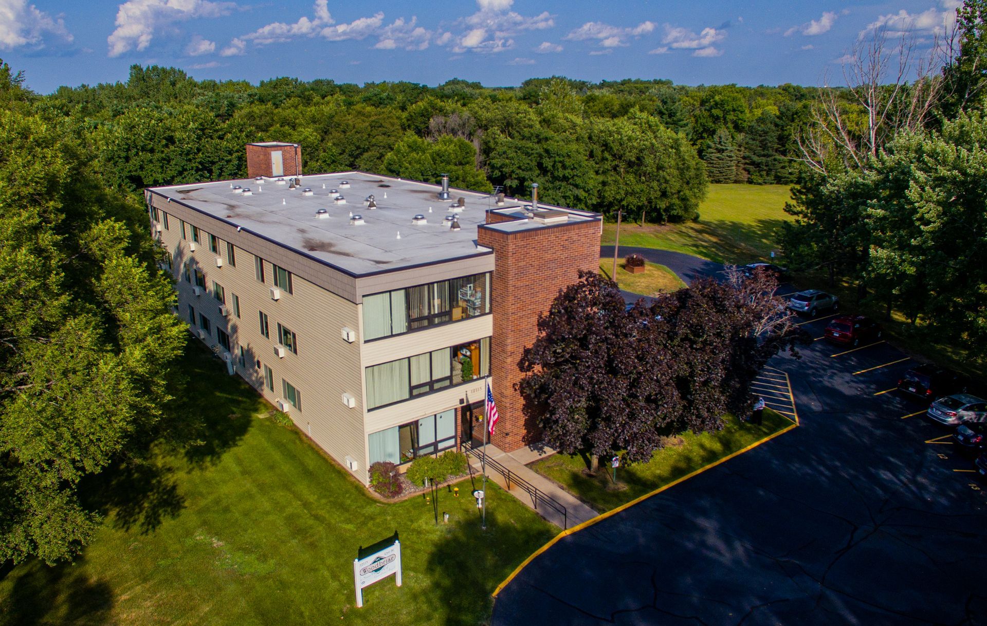 An aerial view of a three-story, beige apartment building with a brick section, surrounded by trees and a parking lot.