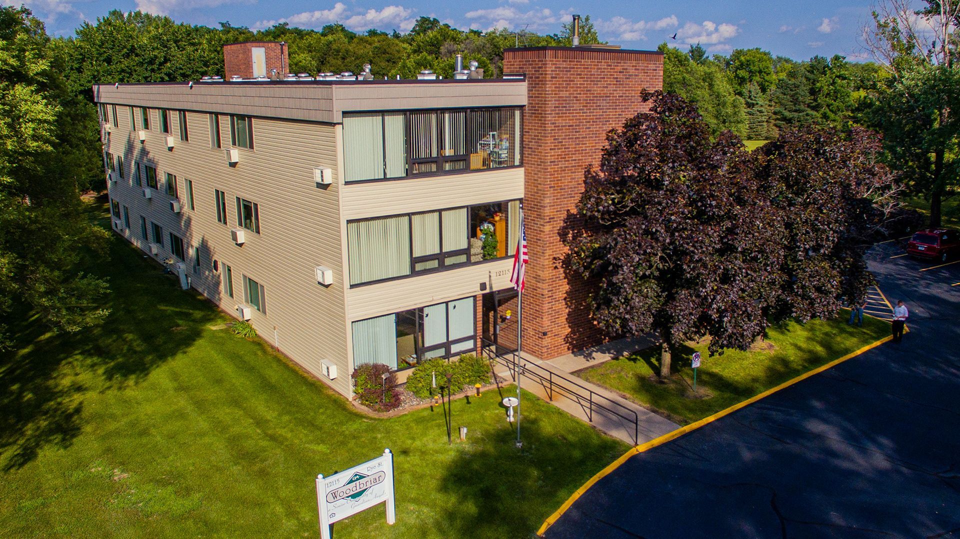A three-story beige apartment building with a brick stairwell section, surrounded by green lawns and trees.