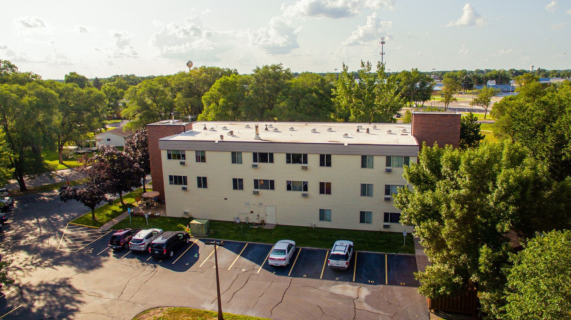 An aerial view of a three-story, light-colored apartment building surrounded by trees with a parking lot in front.