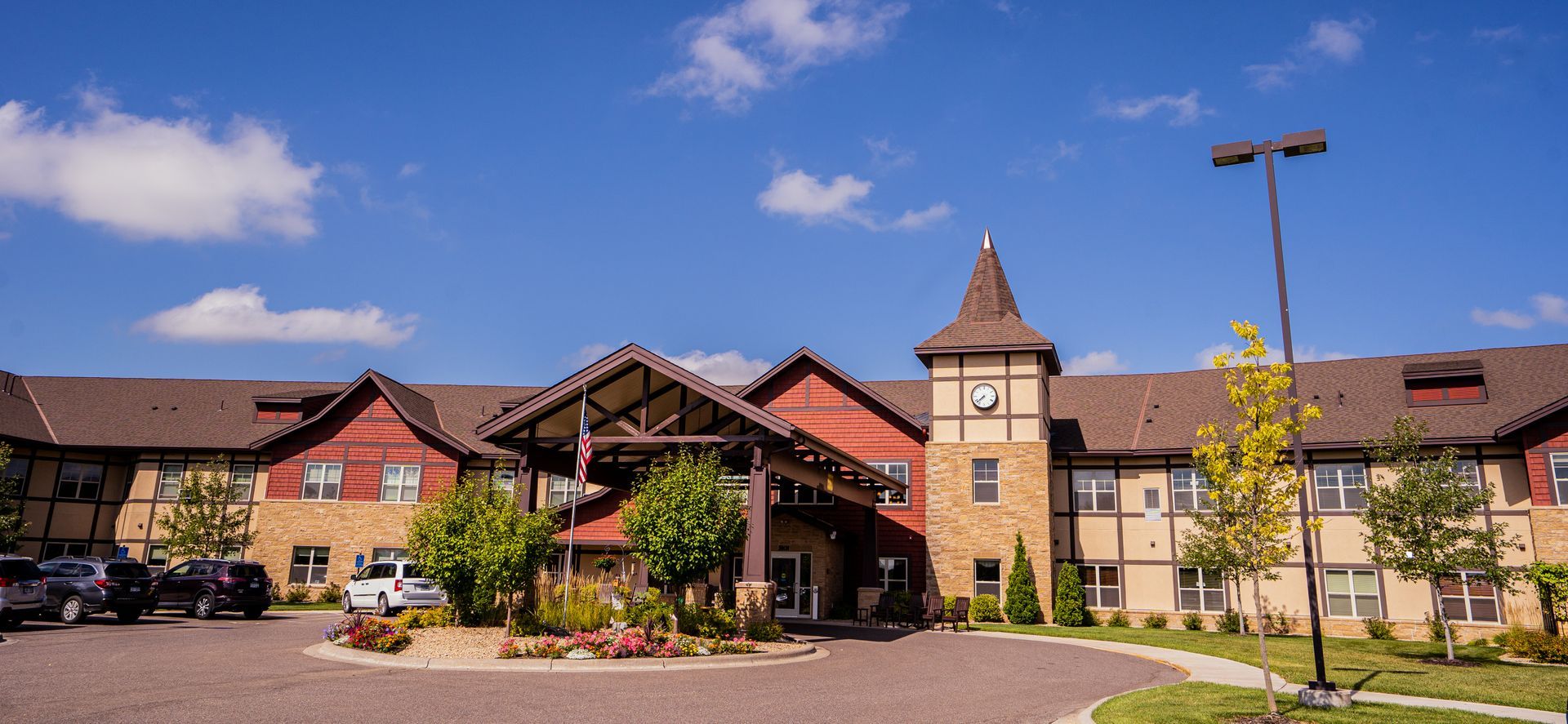 A low-rise hotel with a timber-frame facade and clock tower entrance under a blue sky, with parked cars in the foreground.