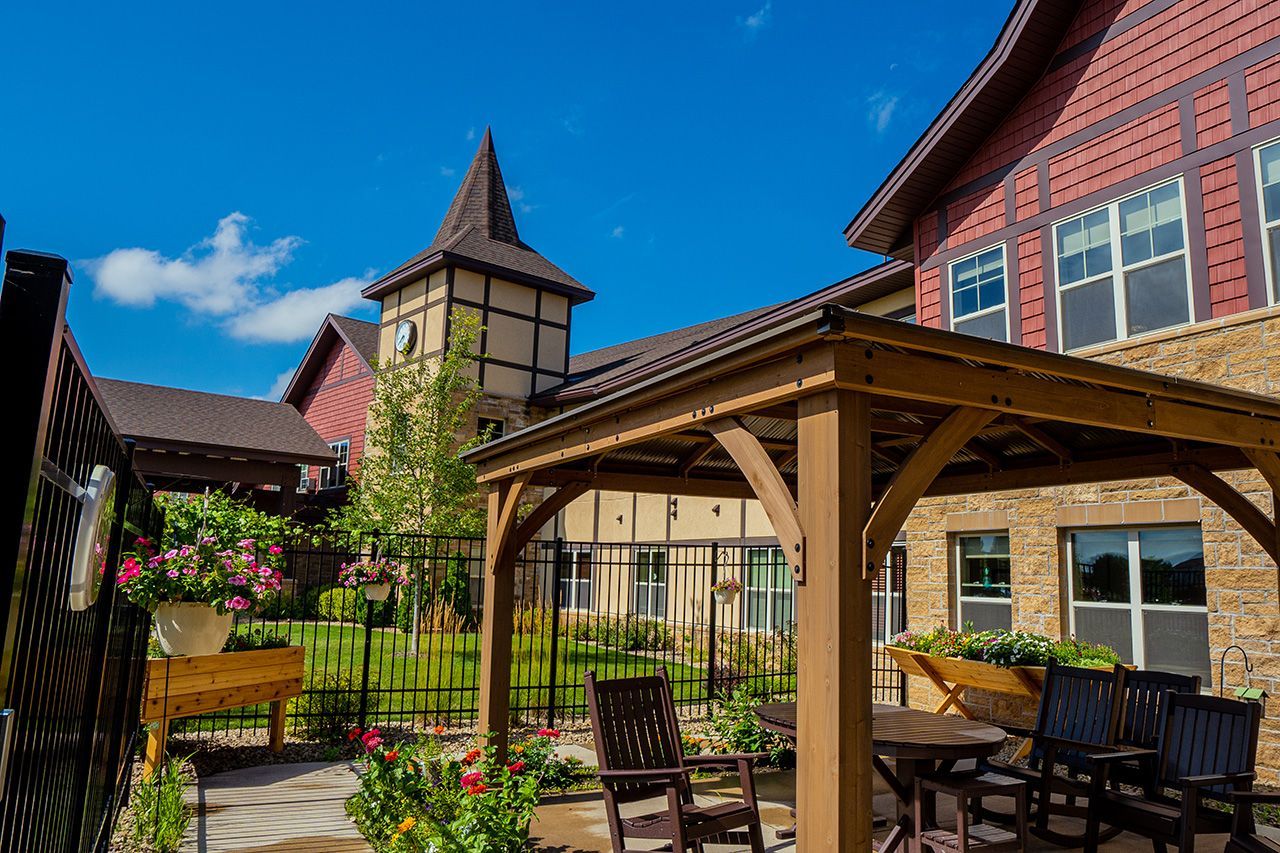 A wooden gazebo with chairs sits in a garden courtyard in front of a multi-story, European-style building under blue sky.