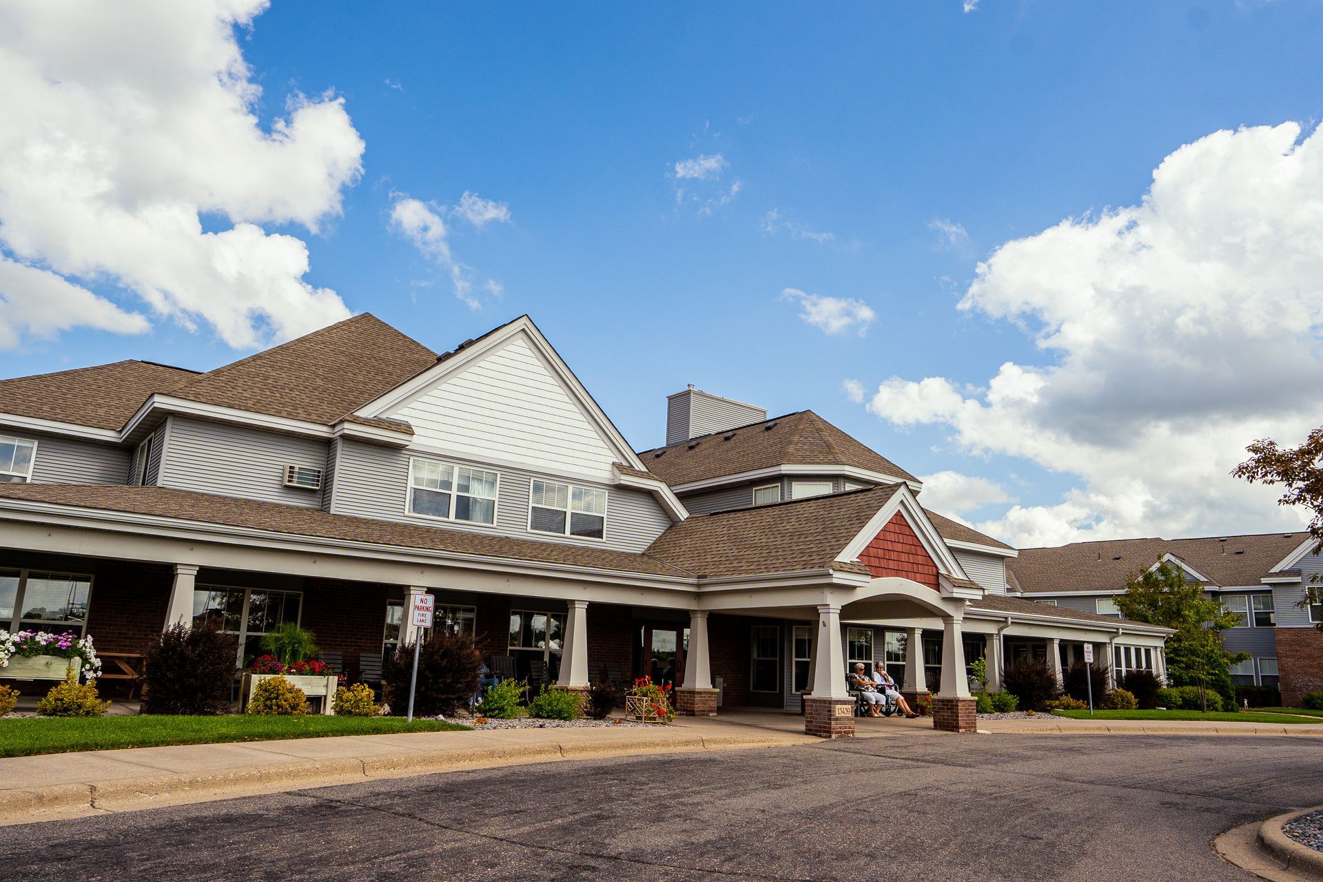 A multi-story building with a brown shingled roof, grey siding, and a covered front porch, set under a blue cloudy sky.