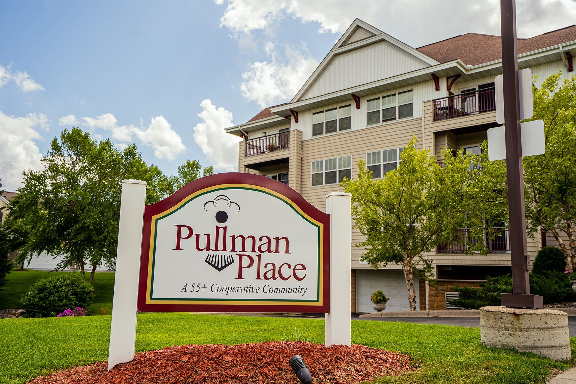 A sign reading "Pullman Place, a 55+ Corporate Community" stands on a grassy lawn in front of a multi-story apartment building.