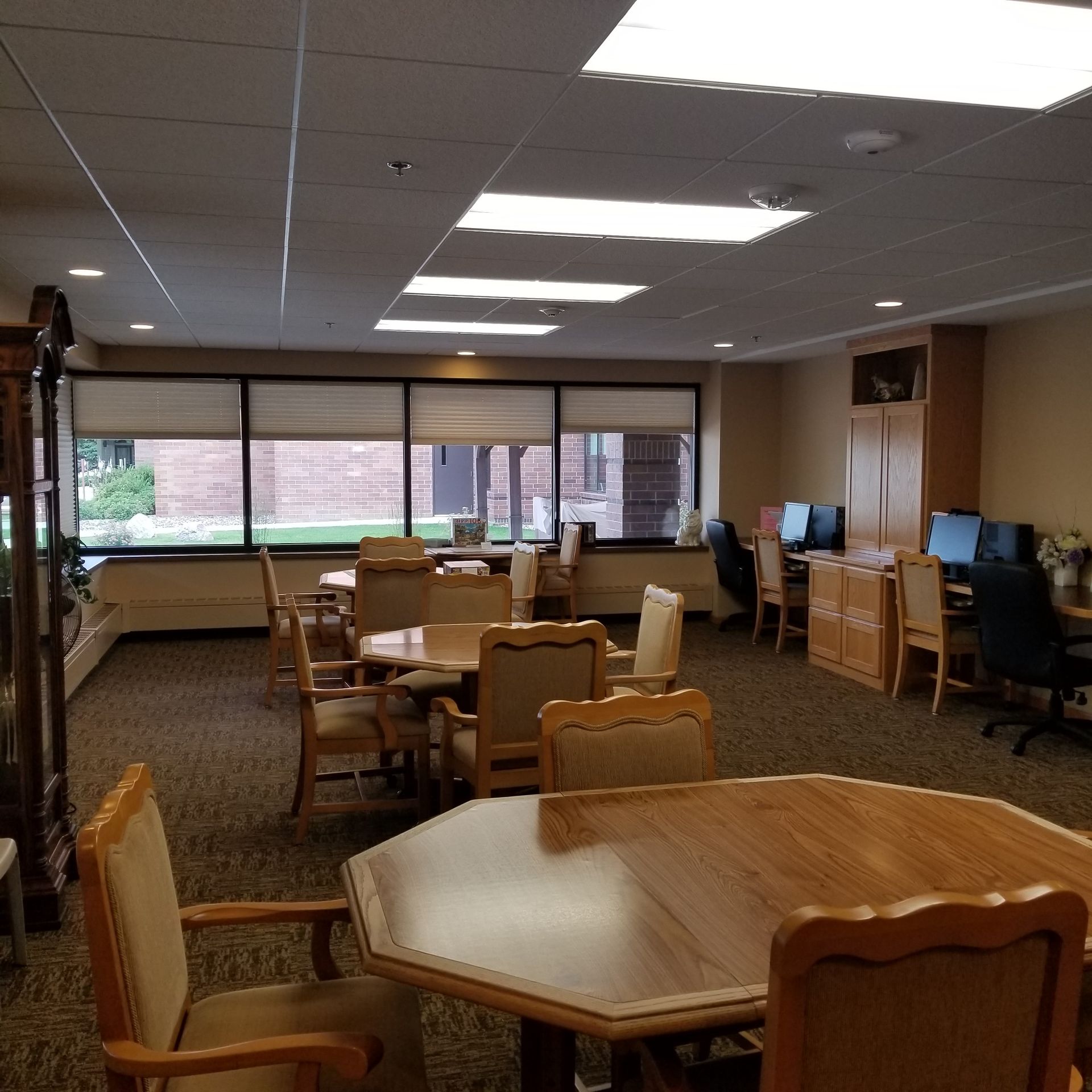 A bright, carpeted room featuring several wooden dining tables with chairs, large windows, and a computer desk area.