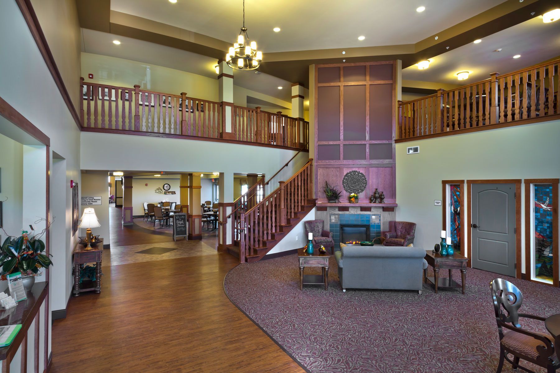 The spacious, multi-level lobby of a senior living facility featuring a central fireplace, staircase, and seating area.
