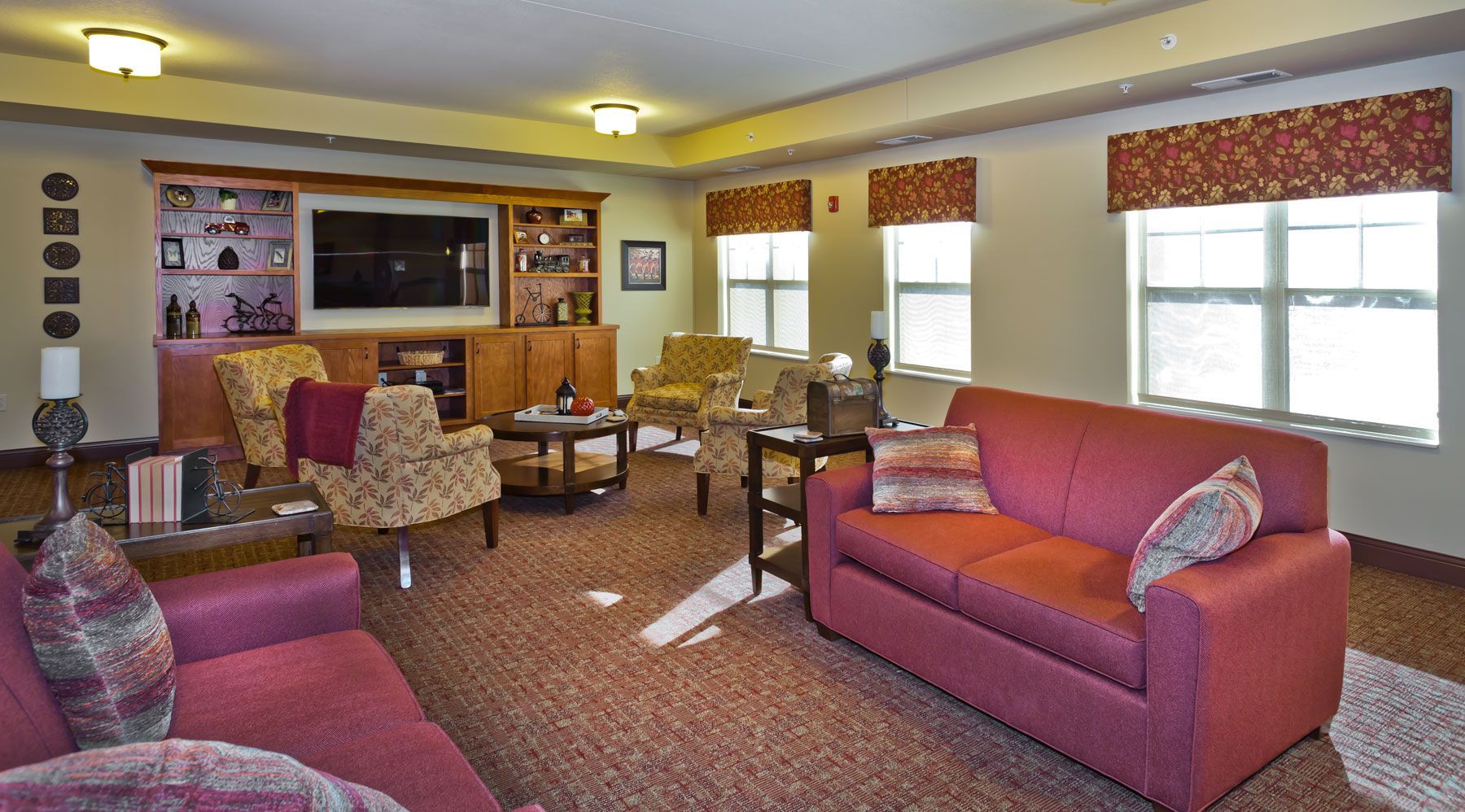 A furnished lounge area featuring a large TV, two red sofas, patterned armchairs, and windows with floral valances.