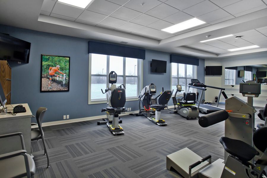A brightly lit fitness center with grey carpet, blue walls, multiple exercise machines, and windows with outdoor views.