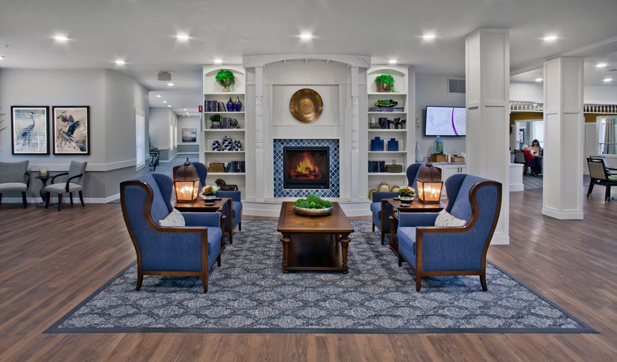A hotel lobby seating area with a central fireplace, two blue armchairs on a patterned rug, and surrounding decor.