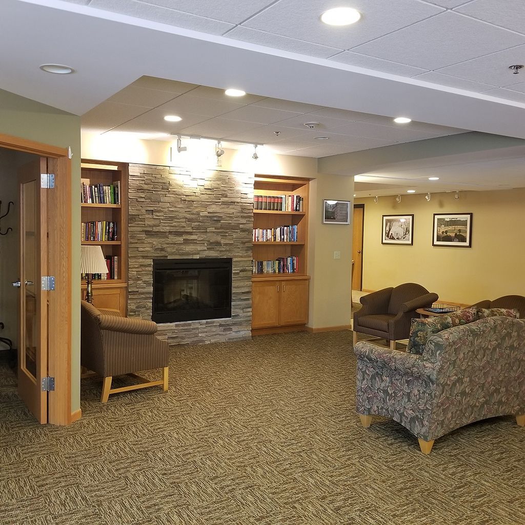 A cozy lounge with a stone fireplace, bookshelves, plush armchairs, and a floral patterned sofa on a carpeted floor.