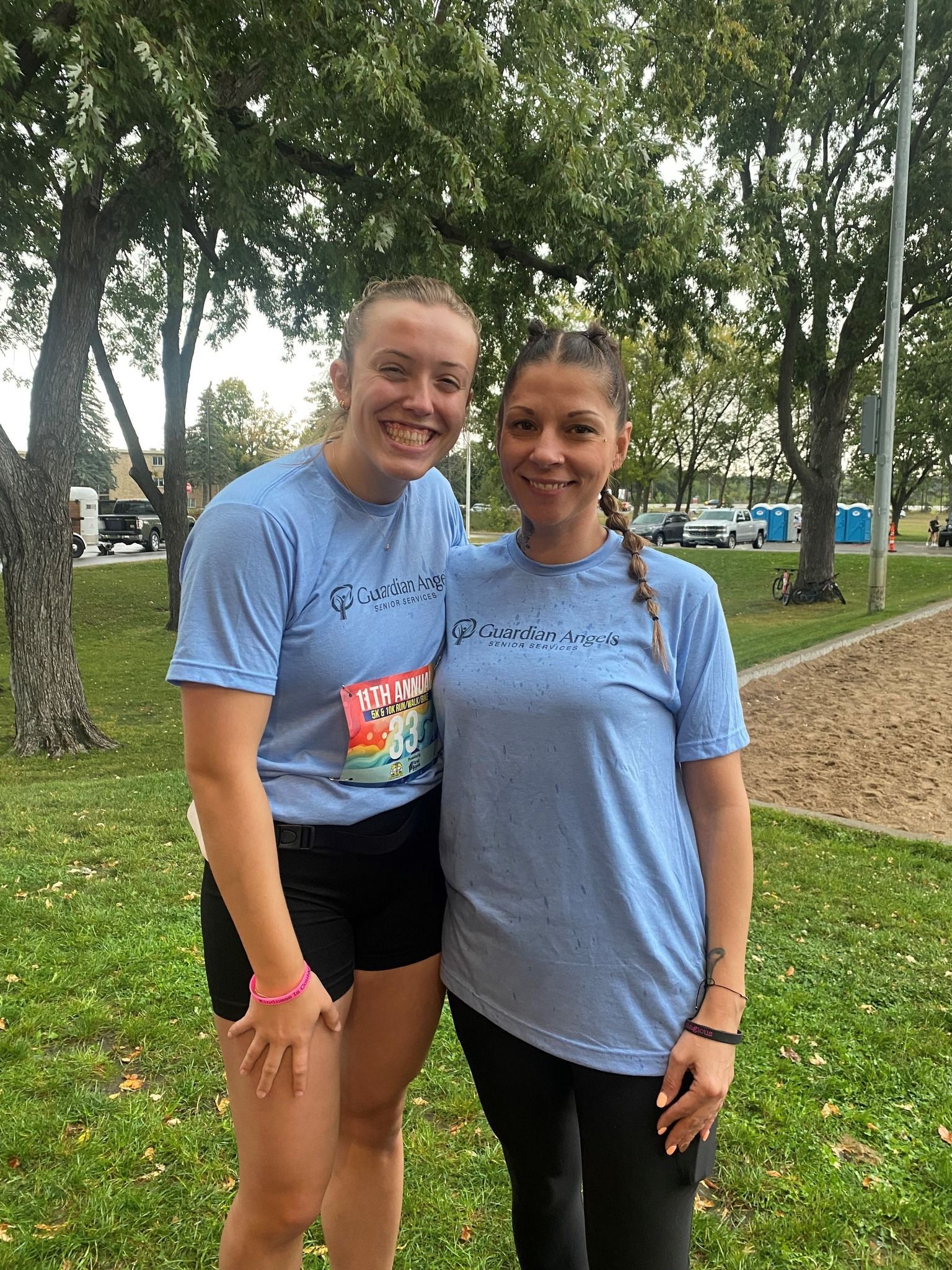 Two people in light blue t-shirts stand side-by-side in a grassy park, smiling at the camera.