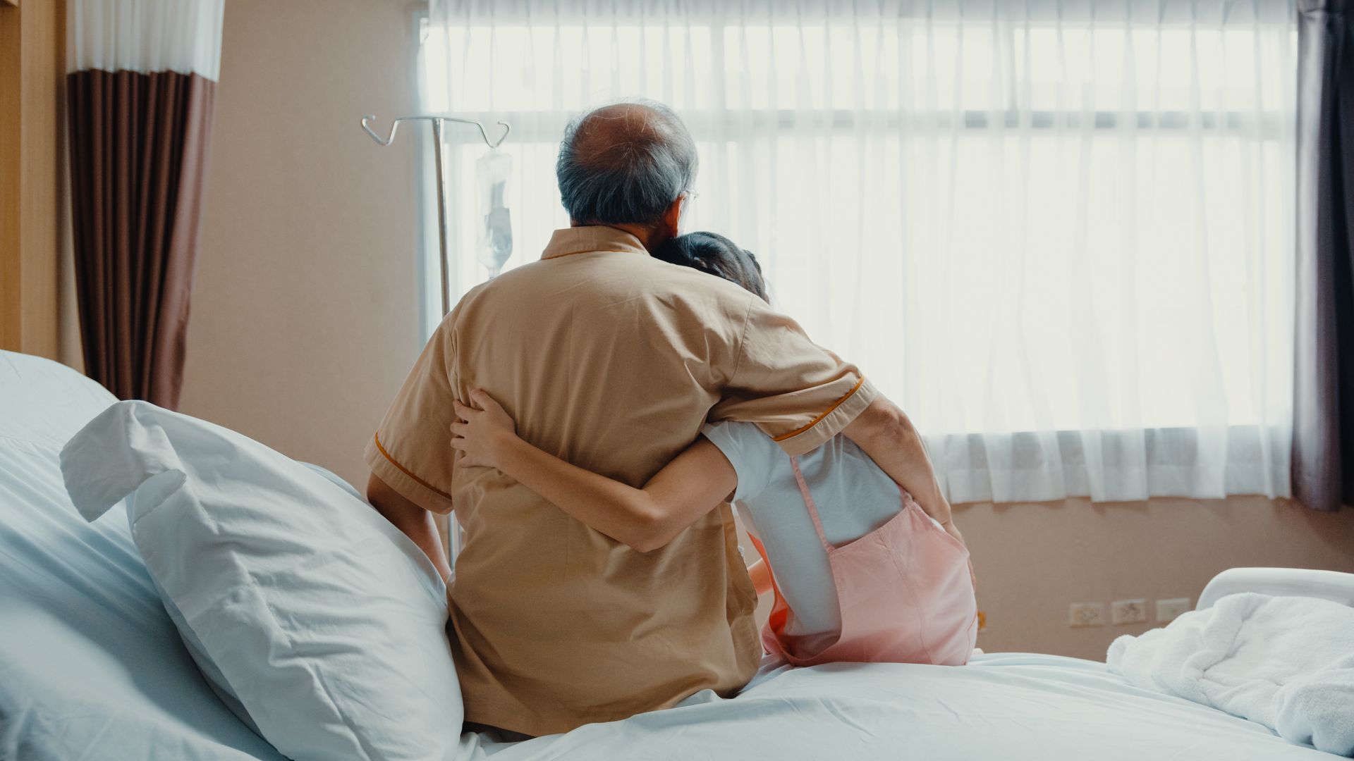 Two people sitting on a hospital bed, viewed from behind, embracing while looking out a window.