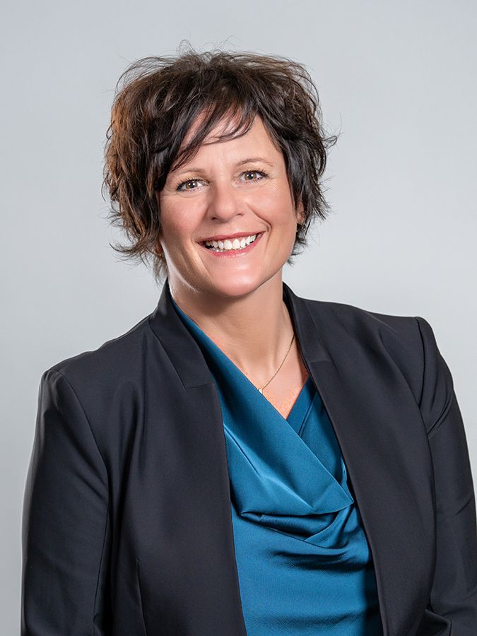 Professional portrait of a person with short brown hair smiling, wearing a dark blue blazer over a teal blouse.