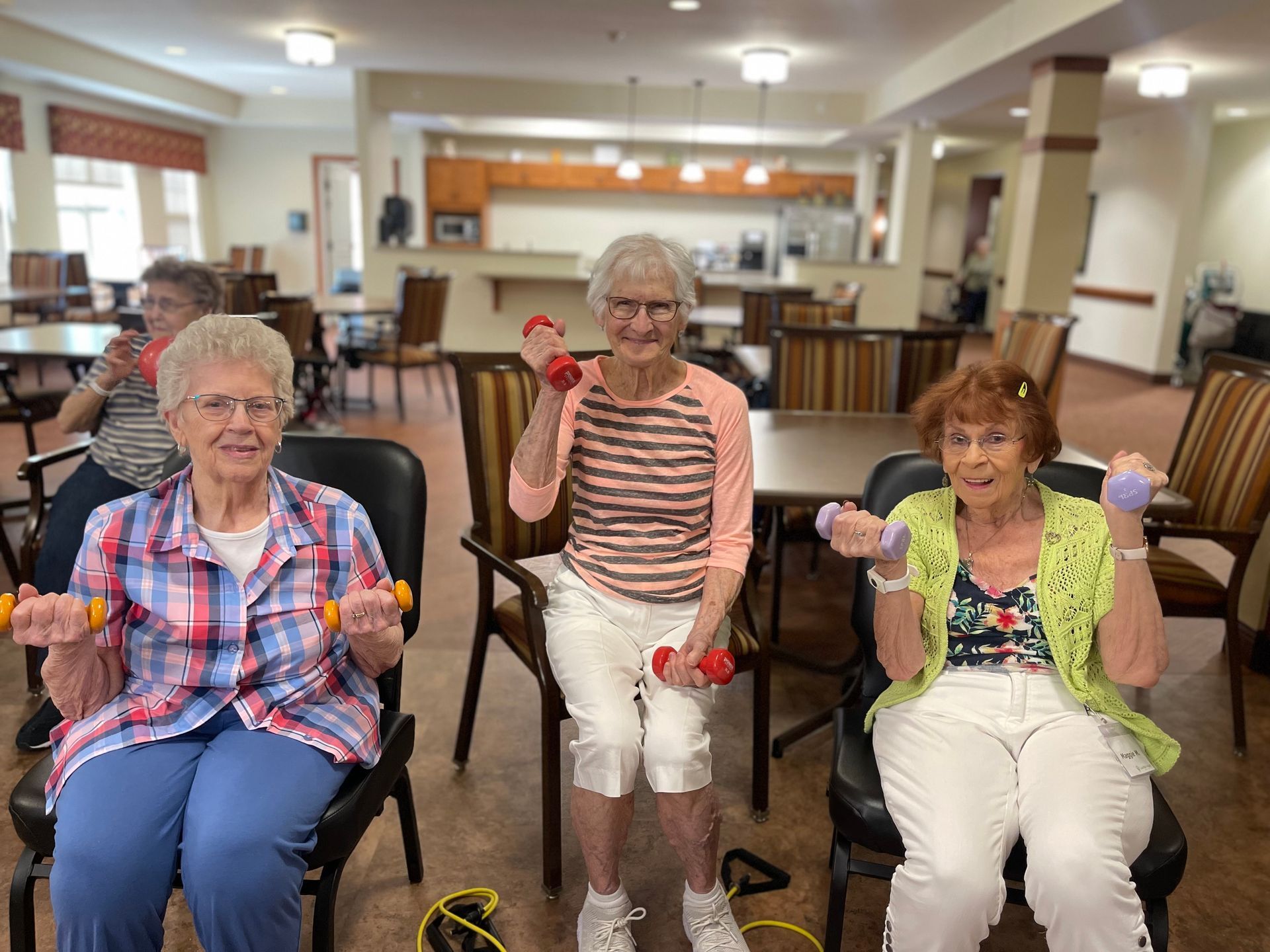 Three people sitting on chairs in a bright room, smiling while lifting small hand weights for an indoor exercise class.