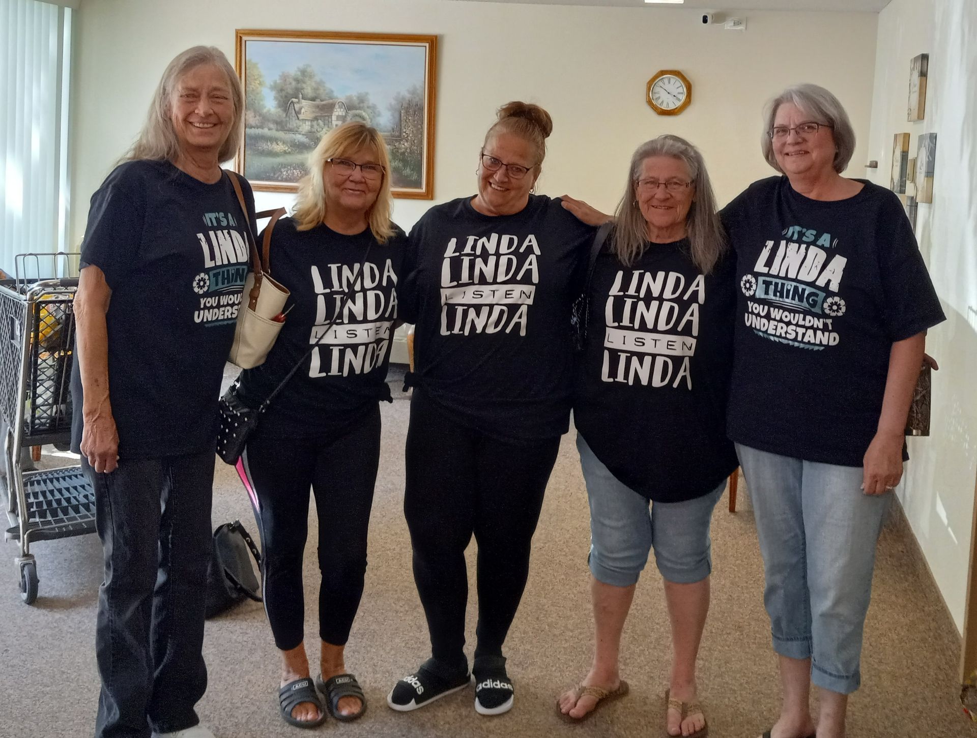 Five smiling people stand in a line indoors, wearing matching dark blue t-shirts with 