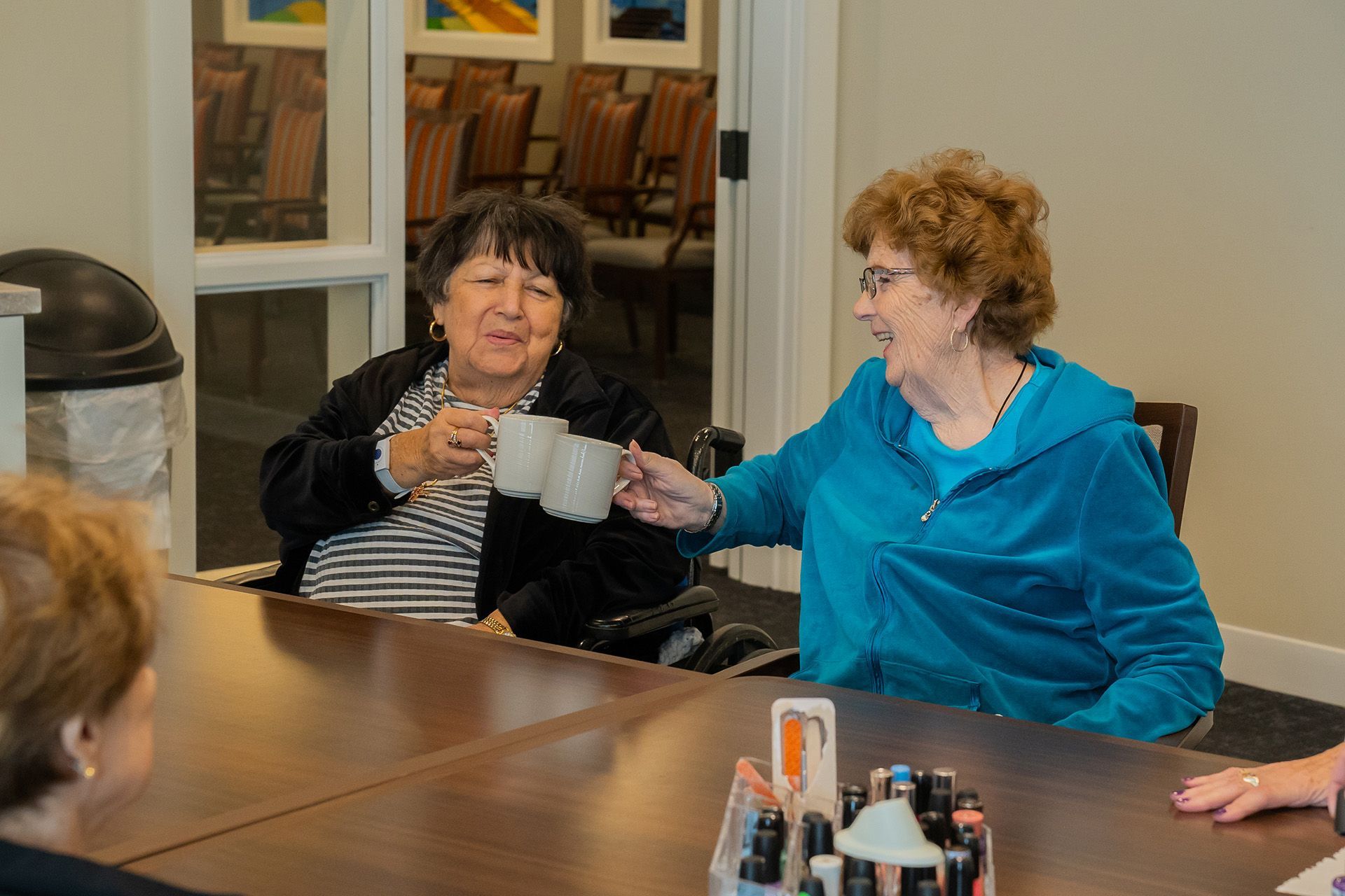 Two people sit at a table, smiling while holding up mugs in a toast, with others present in a well-lit indoor setting.